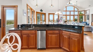 A spacious kitchen with wooden cabinets, a stainless steel dishwasher, and large windows offering a view of the outdoors. - Wellfleet: Soundfront | Pool and Hot Tub | Dogs OK (Corolla)