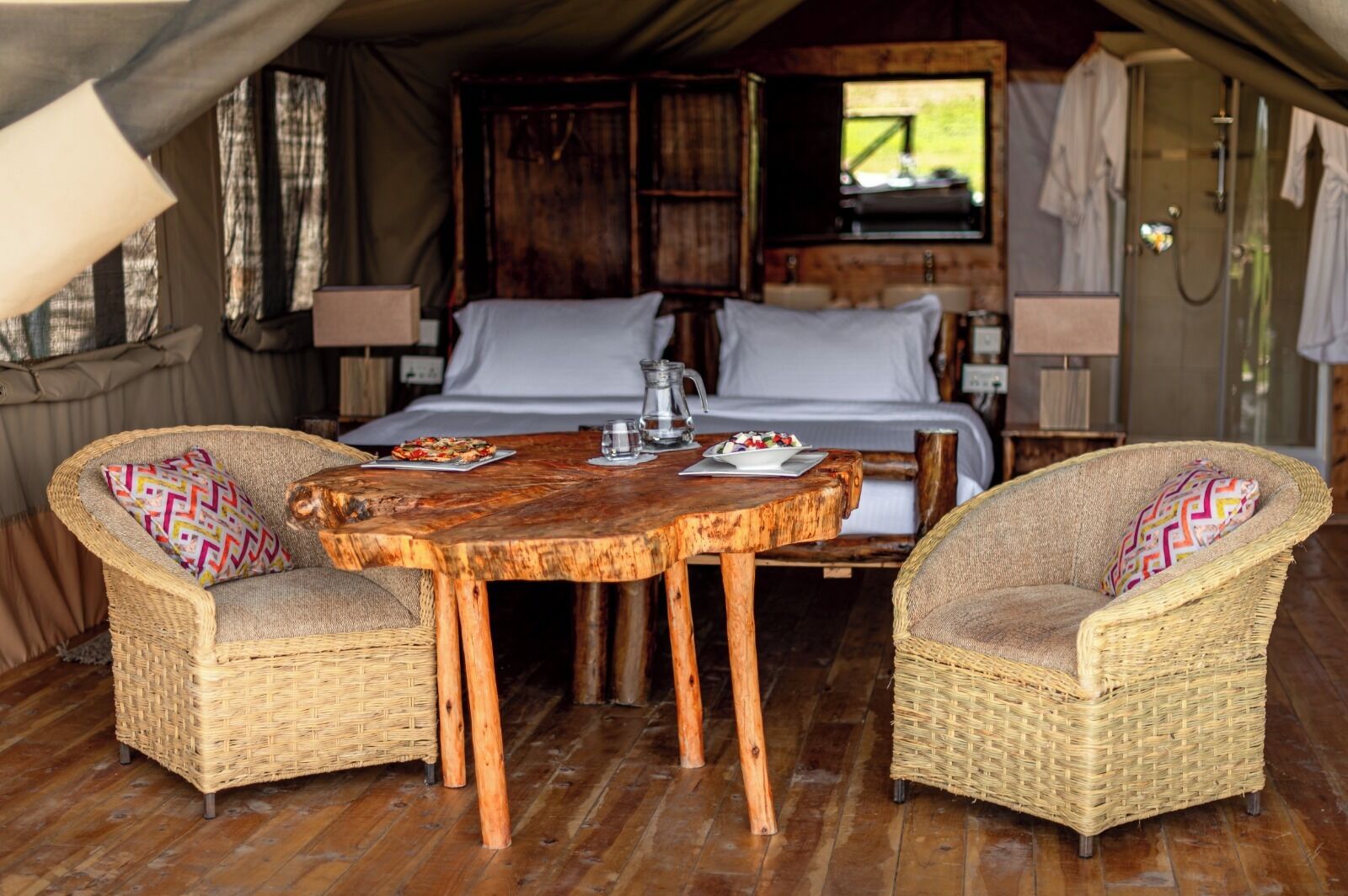 Een gezellig tentverblijf met een bed, twee rieten stoelen, een houten tafel en een kleine kitchenette.