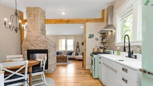 A kitchen with a stone fireplace, wooden beams, a dining area with a table and chairs, and a view of the living room.