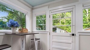 A room with a white door, a window with a view of greenery, a table with a chair, and a vase with flowers.