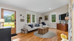 A spacious living room with a wooden floor, a sofa set, a coffee table, and a TV mounted on the wall.