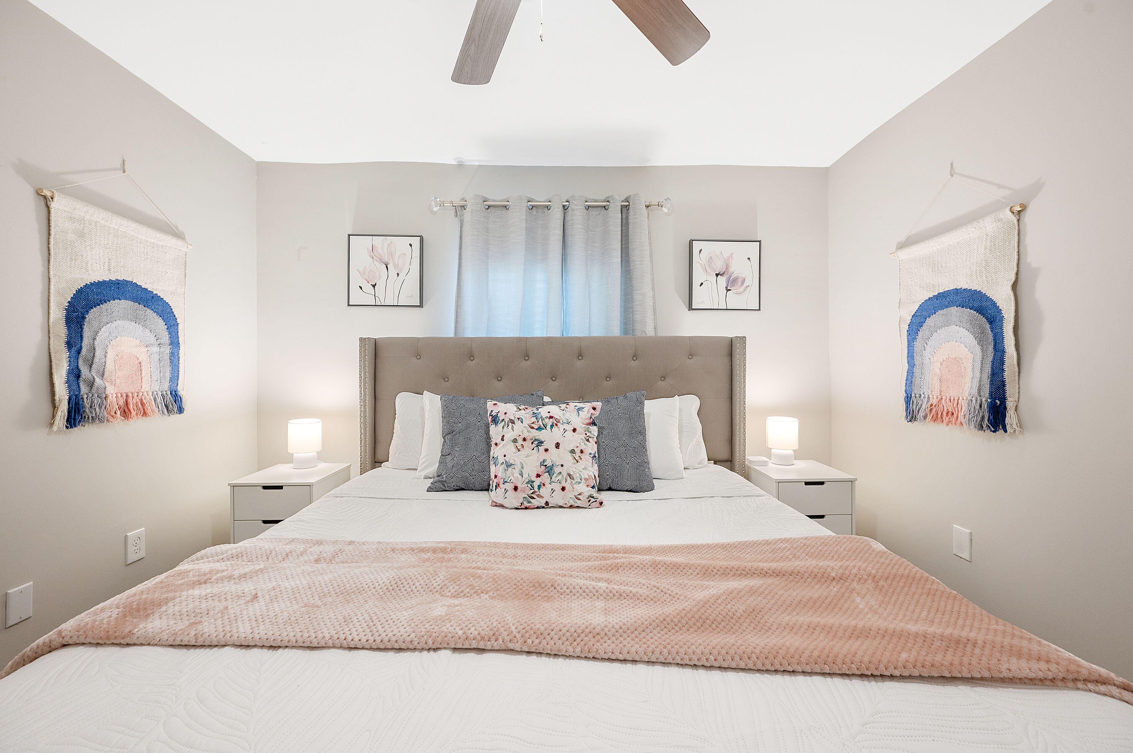 Une chambre à coucher avec un grand lit, deux tables de chevet avec des lampes, un ventilateur de plafond et deux tapisseries murales.