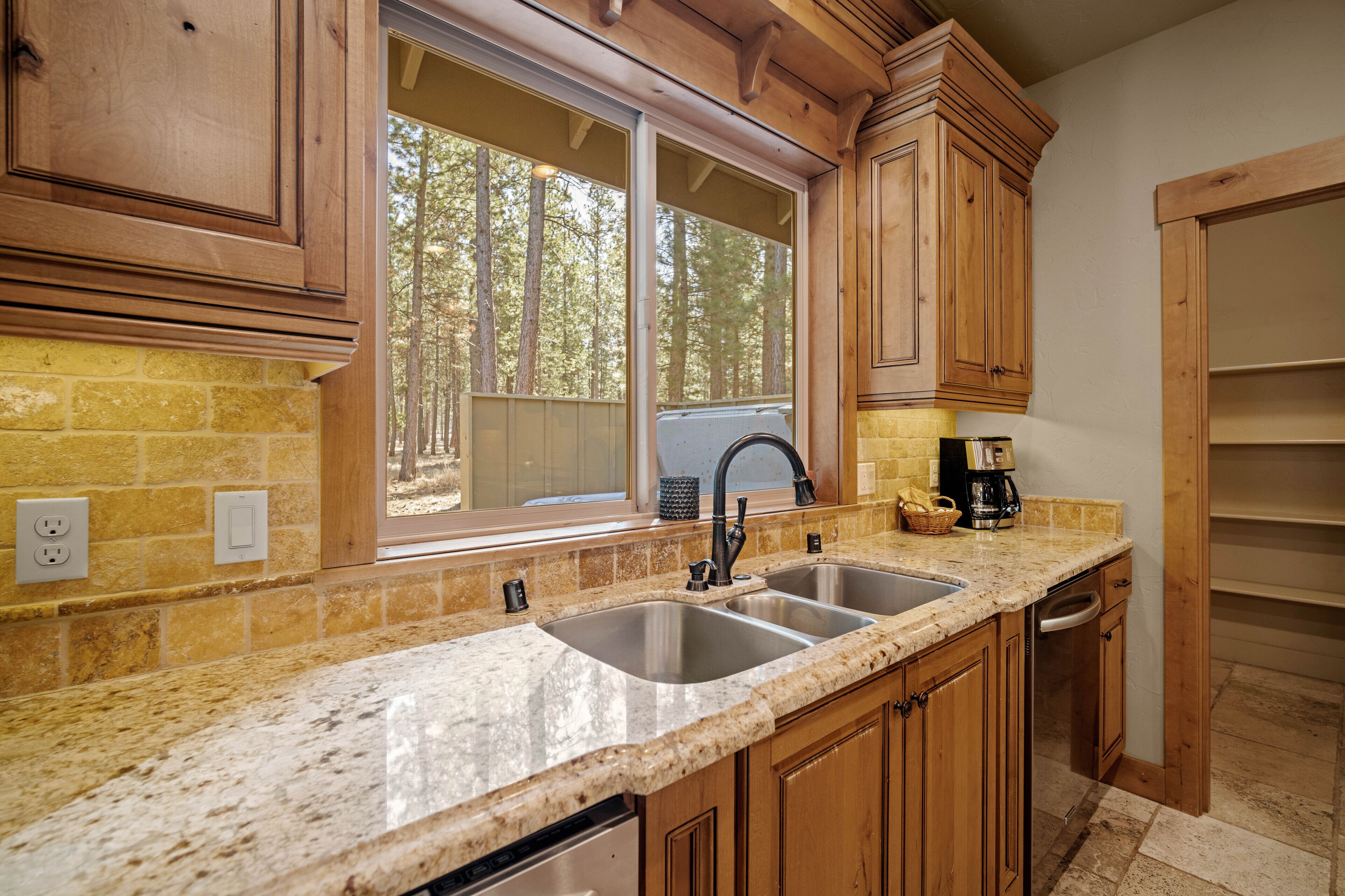 A kitchen with granite countertops, wooden cabinets, a double sink, and a window with a view of trees.
