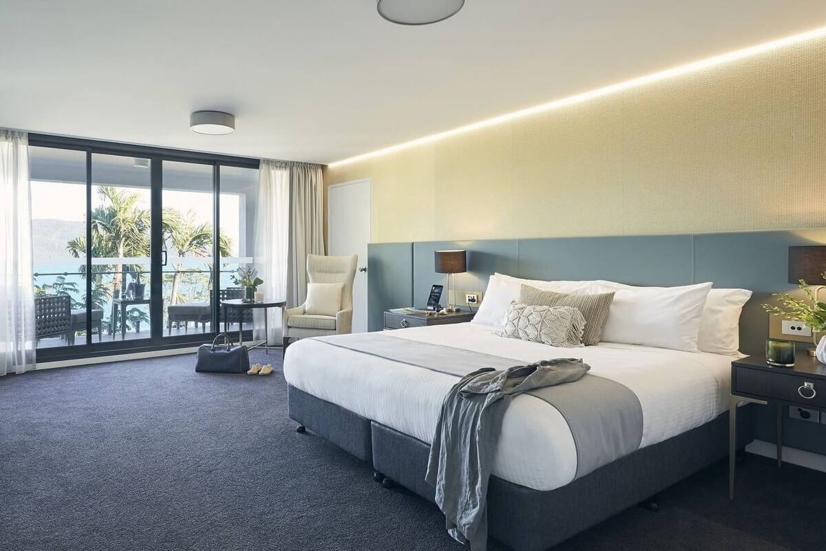 A modern hotel room with a large bed, a nightstand, a chair, and a view of the outdoors through large windows.