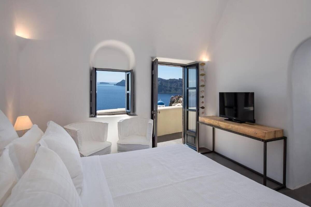 A hotel room with a bed, a television on a wooden console, a seating area with a chair, and a balcony with a view of the sea.