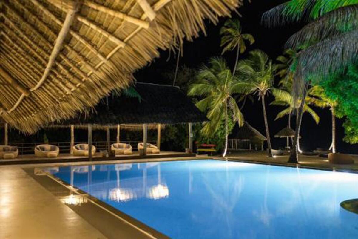 A swimming pool with lounge chairs under a thatched roof, surrounded by palm trees.