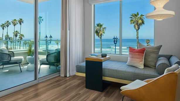 Une chambre moderne dotée d’une grande fenêtre donnant sur la plage, d’un canapé avec des coussins et d’une petite table sur laquelle est posé un livre.