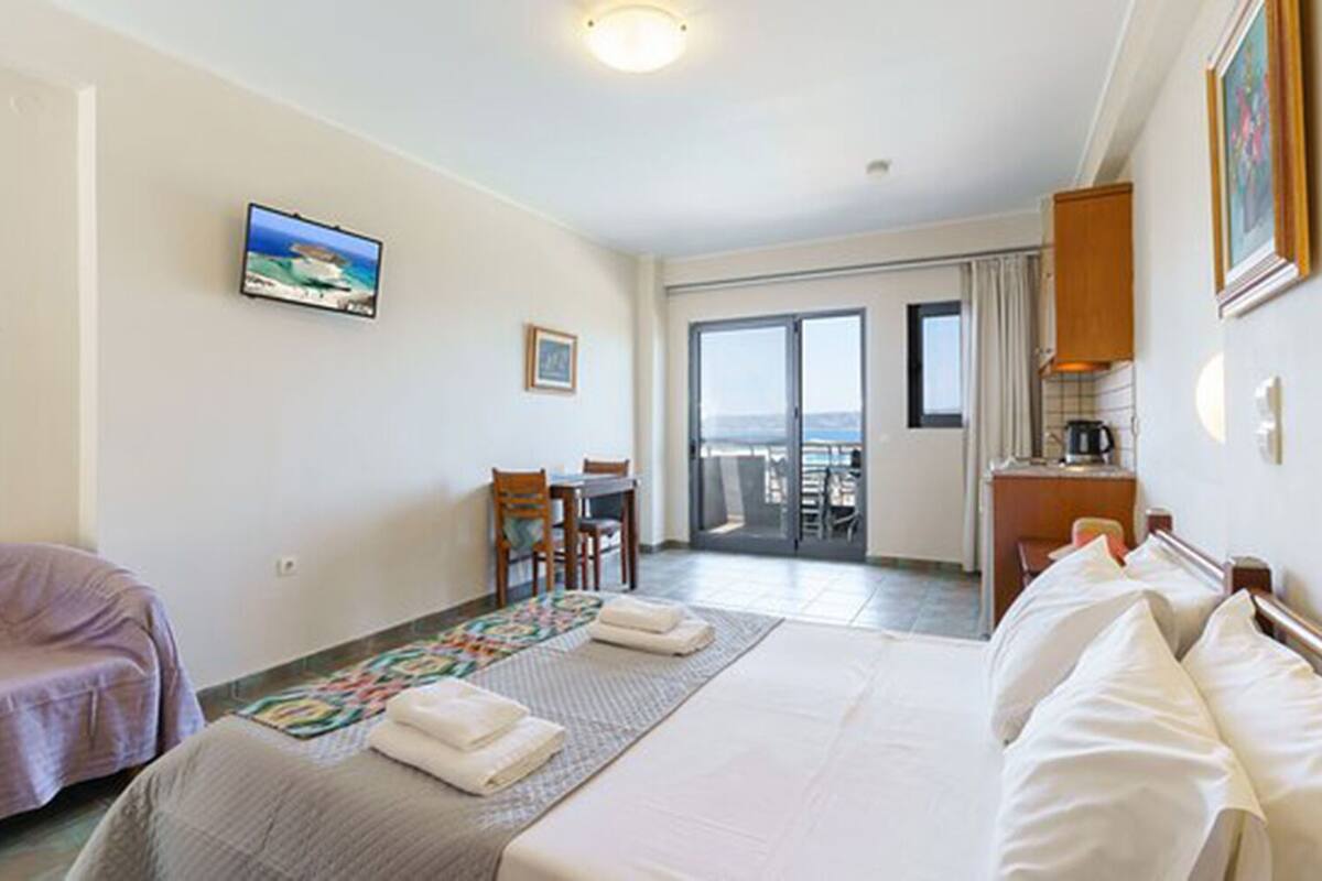 A hotel room with a bed, a desk, a chair, a small table, and a view of the sea through a sliding door.