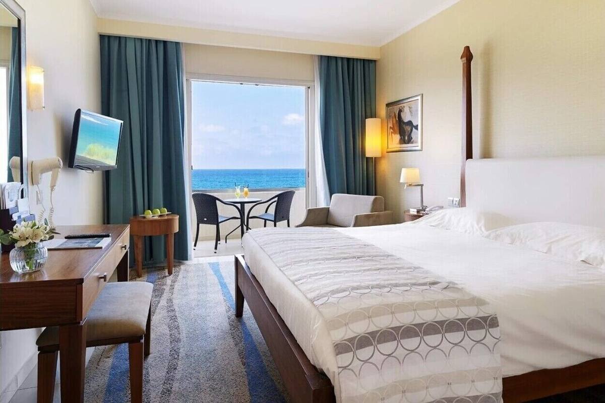 A hotel room with a large bed, a desk, a chair, a small table with drinks, a TV, and a view of the ocean.