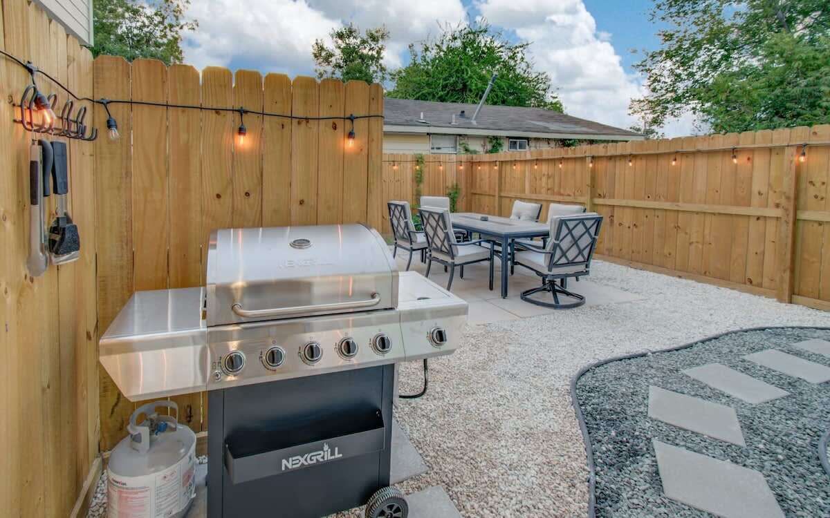 A backyard with a NEGRILL gas grill, a wooden fence, a patio table with chairs, and a stone-patterned rug.