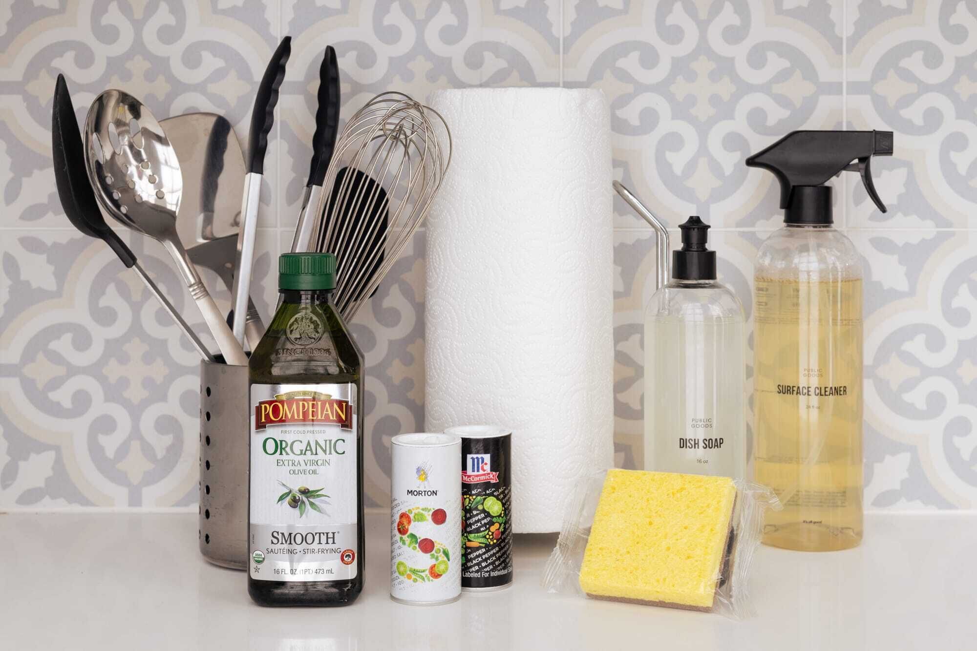 A kitchen counter with various items including a bottle of organic extra virgin olive oil, a can of Morton seasoning, a sponge, a dish soap bottle, and a roll of paper towels.