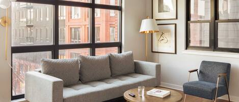 A modern living room with a grey sofa, a round coffee table, and a blue armchair. There are large windows with black frames, a floor lamp, and framed artwork on the wall.