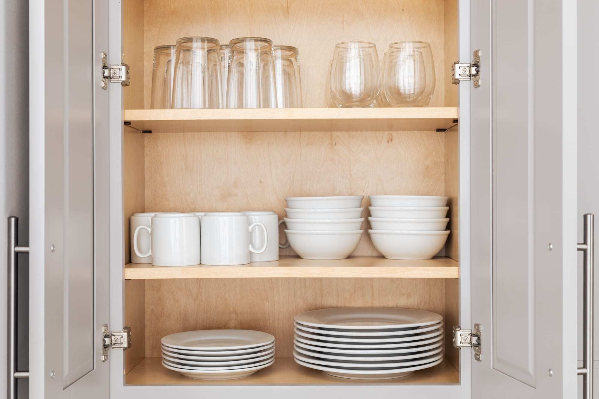 Un aparador con tres repisas: la repisa superior contiene frascos de vidrio, la del medio tazas y recipientes blancos, y la inferior platos apilados.
