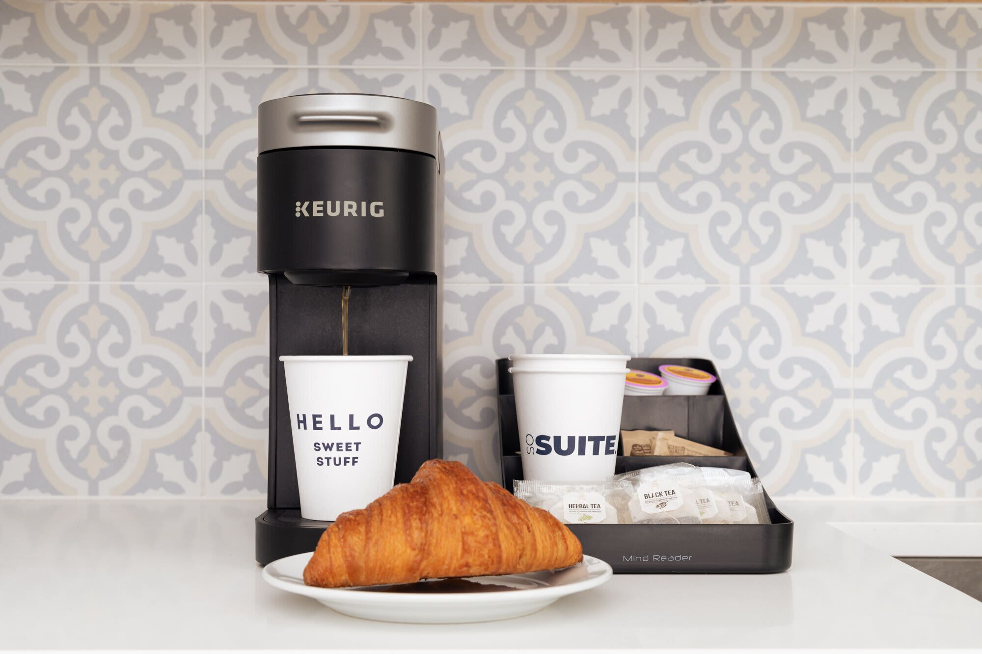 Una cafetera Keurig, una taza Hello Sweet Stuff, una taza de suite, un Mind Reader y un croissant en un plato.