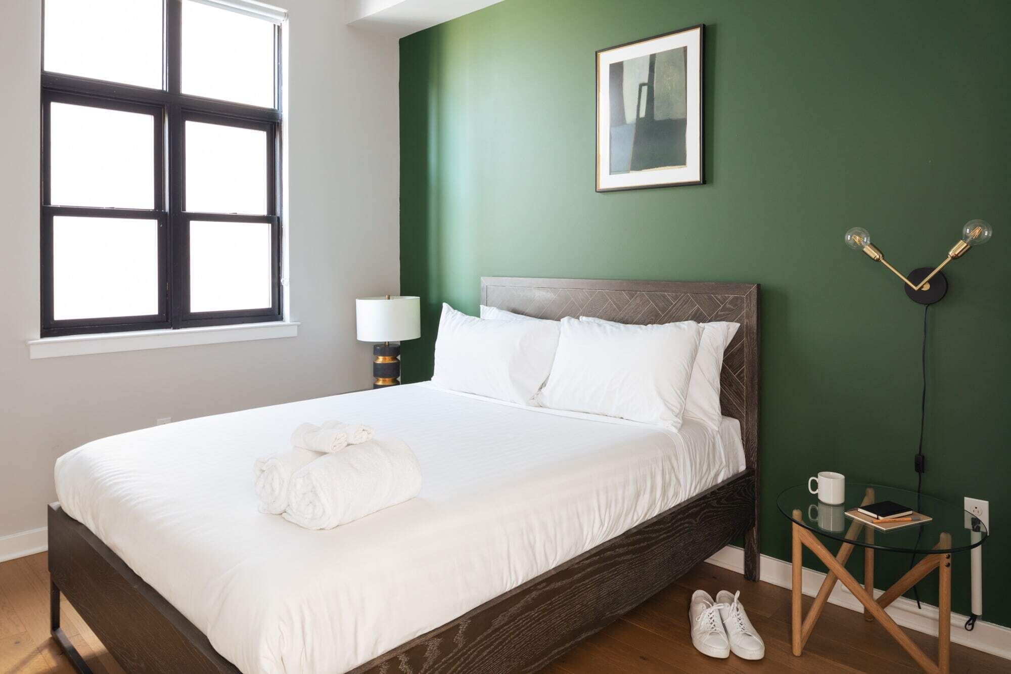Una cama bien hecha con sábanas blancas y una toalla enrollada, una lámpara de noche, un cuadro en la pared y una mesita con un libro y una taza.