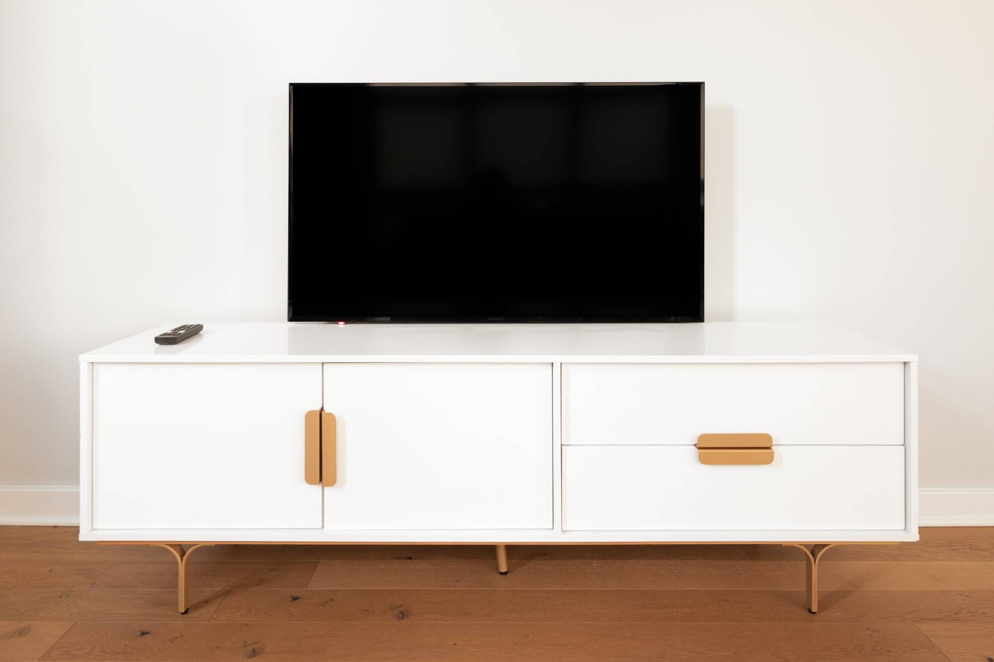 A white TV stand with a flat-screen TV mounted above it, a remote control on the left, and two drawers with wooden handles.