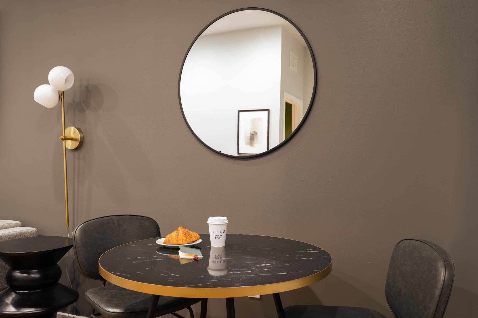 A round table with a black marble top, a plate with a croissant, a coffee cup, and a book. There are two black chairs around the table. A gold floor lamp with three white lampshades is to the left. A large oval mirror hangs on the wall above the table.
