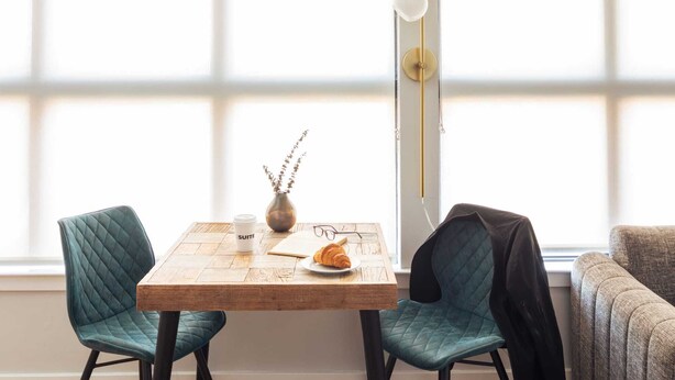 En matplats med ett träbord, två turkosa stolar, en vas med torkade blommor, en croissant och en kaffekopp märkt med texten "SUITE".