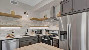 A modern kitchen with a stainless steel refrigerator, a microwave, a stove, and a dishwasher. There are wooden shelves above the counter and a light fixture hanging from the ceiling. - The Grove Estate 8BD/7BA!  Perfect For Groups! (Fredericksburg)