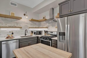 A modern kitchen with a stainless steel refrigerator, a microwave, a stove, and a dishwasher. There are wooden shelves above the counter and a light fixture hanging from the ceiling. - The Grove Estate 8BD/7BA!  Perfect For Groups! (Fredericksburg)