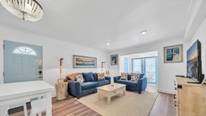 Un salon moderne avec deux canapés, une table basse et un téléviseur. On y trouve un buffet en bois et une table blanche avec des chaises. Le sol est recouvert de parquet et la pièce est dotée d’une grande fenêtre avec des stores.