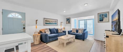 A modern living room with two sofas, a coffee table, and a TV. There is a wooden sideboard and a white table with chairs. The room has wooden flooring and a large window with blinds.