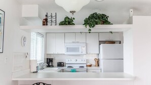 A modern kitchen with white cabinets, a microwave, a stove, and a refrigerator. There are two plants on the countertop and a hanging light fixture above. - 2nd floor beach condo (Cocoa Beach)
