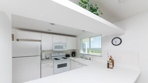 Une cuisine moderne avec des meubles blancs, un four à micro-ondes, une cuisinière et un évier. Il y a une fenêtre avec des stores, une horloge au mur et une plante suspendue au plafond.