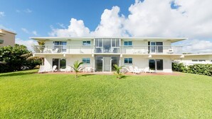 Une maison moderne à deux étages, dotée d’un grand jardin à l’avant, d’un balcon à l’étage supérieur et surplombant un ciel d’un bleu éclatant.