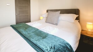 Uma cama bem arrumada com um cobertor verde, uma porta de madeira e uma luminária fixada na parede.