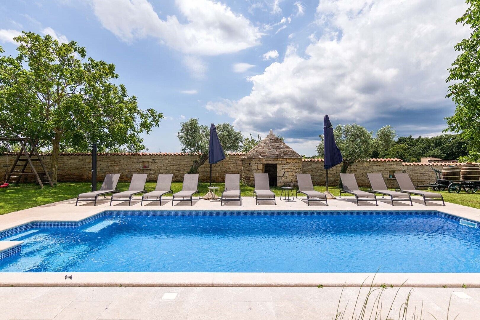 A swimming pool with lounge chairs and umbrellas, surrounded by stone walls and trees.