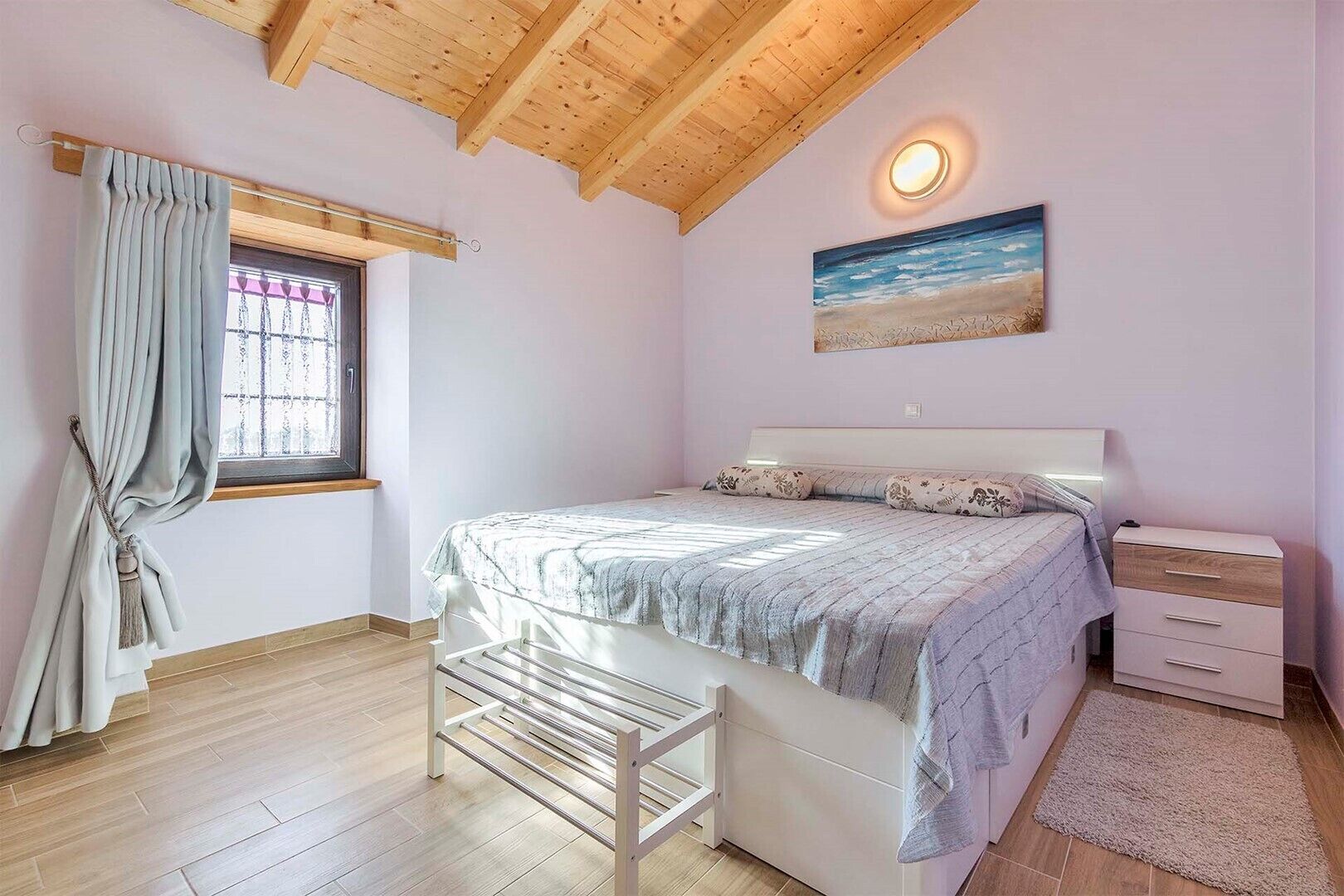 A bedroom with a bed, bedside tables, a window with curtains, a painting, and a wooden ceiling.