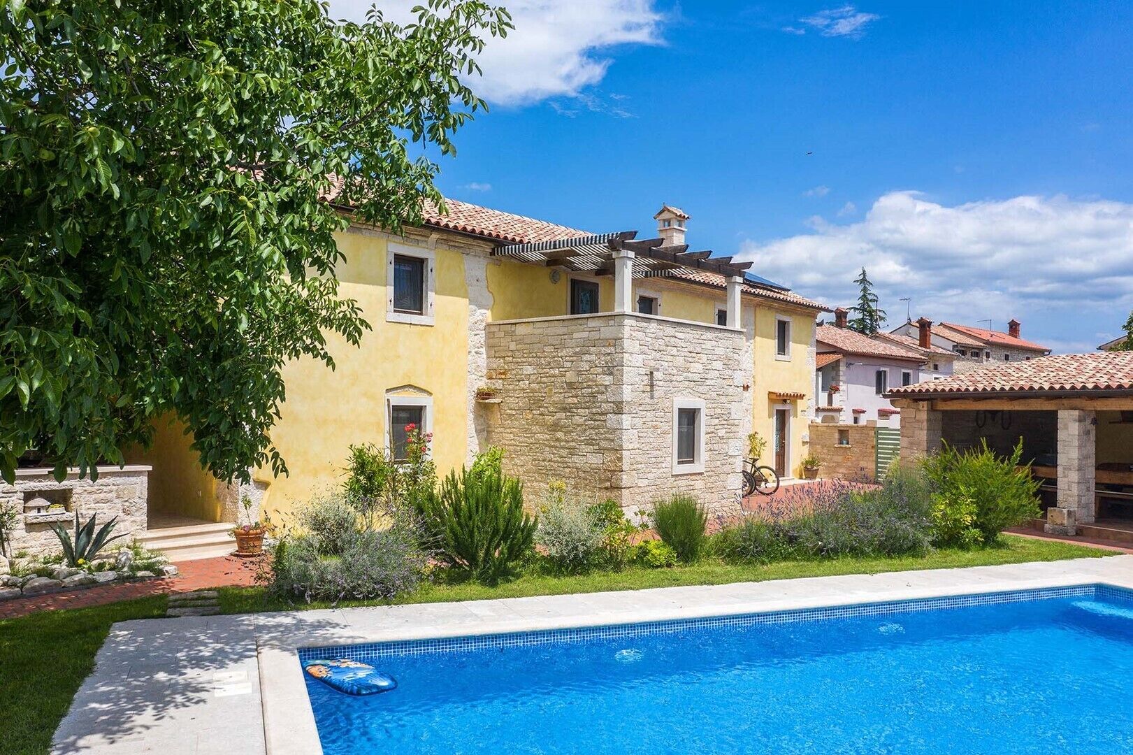 A poolside view of a yellow building with a stone structure, surrounded by greenery and a clear blue sky.