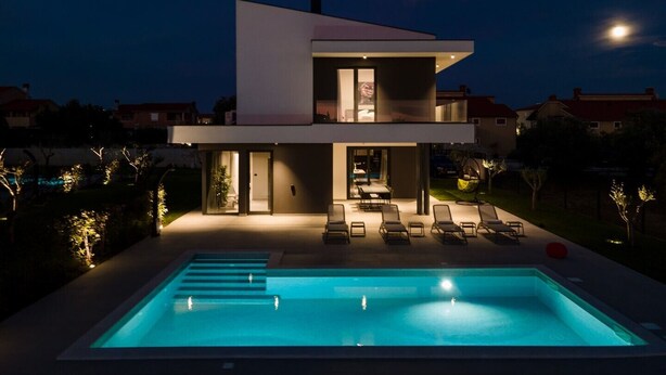 A modern two-story house with a swimming pool, outdoor seating, and a well-lit patio area.