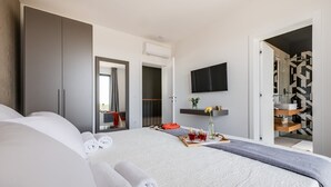 A modern hotel room with a large bed, a flat-screen TV, a small table with a tray of drinks, and a bathroom with a geometric tile backsplash.
