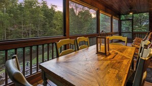 A wooden deck with a table and chairs, large windows offering a view of a forest, and a hanging light fixture. - Bear Shenanigans in Blue Ridge (Blue Ridge)