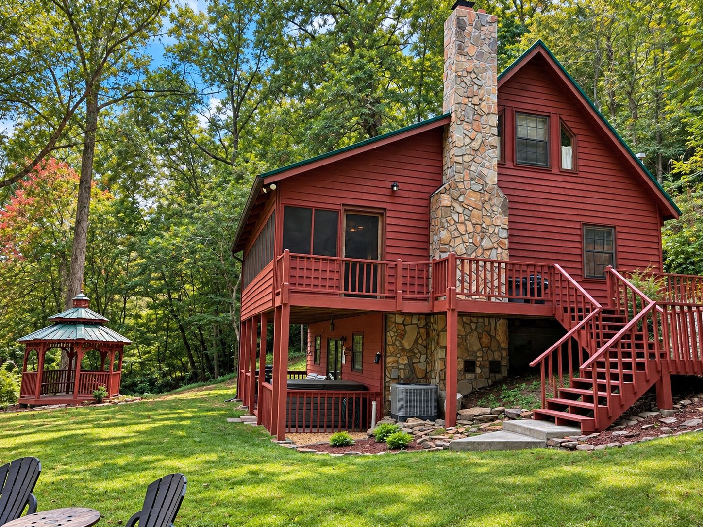 A two-story cabin with a stone chimney, a deck, and a gazebo in a wooded setting.