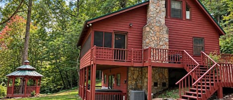 A two-story cabin with a stone chimney, a deck, and a gazebo in a wooded setting.