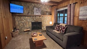 A cozy living room with a stone fireplace, a television mounted on the wall, a sofa, a coffee table, and a window with curtains. - Bigfoot's River Retreat (Mineral Bluff)