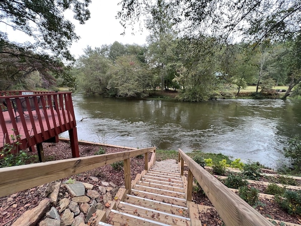 Una escalera de madera que desciende hacia un río, rodeada de árboles y una terraza de madera con sillas.
