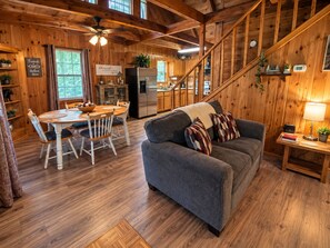 A cozy cabin interior with a dining area, a sofa, and a staircase leading to a loft.