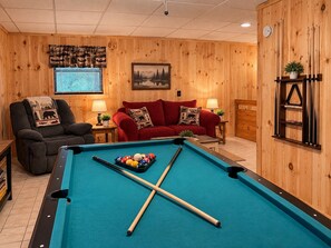 Une table de billard avec des boules et des queues, un canapé rouge, un fauteuil gris et des murs en bois.