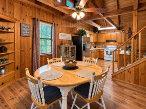Interior de una cabaña de madera con una mesa de comedor, sillas, una zona de cocina y una escalera.