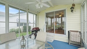 Un patio techado con una mesa de vidrio, sillas, un ventilador de techo y vista al jardín trasero.