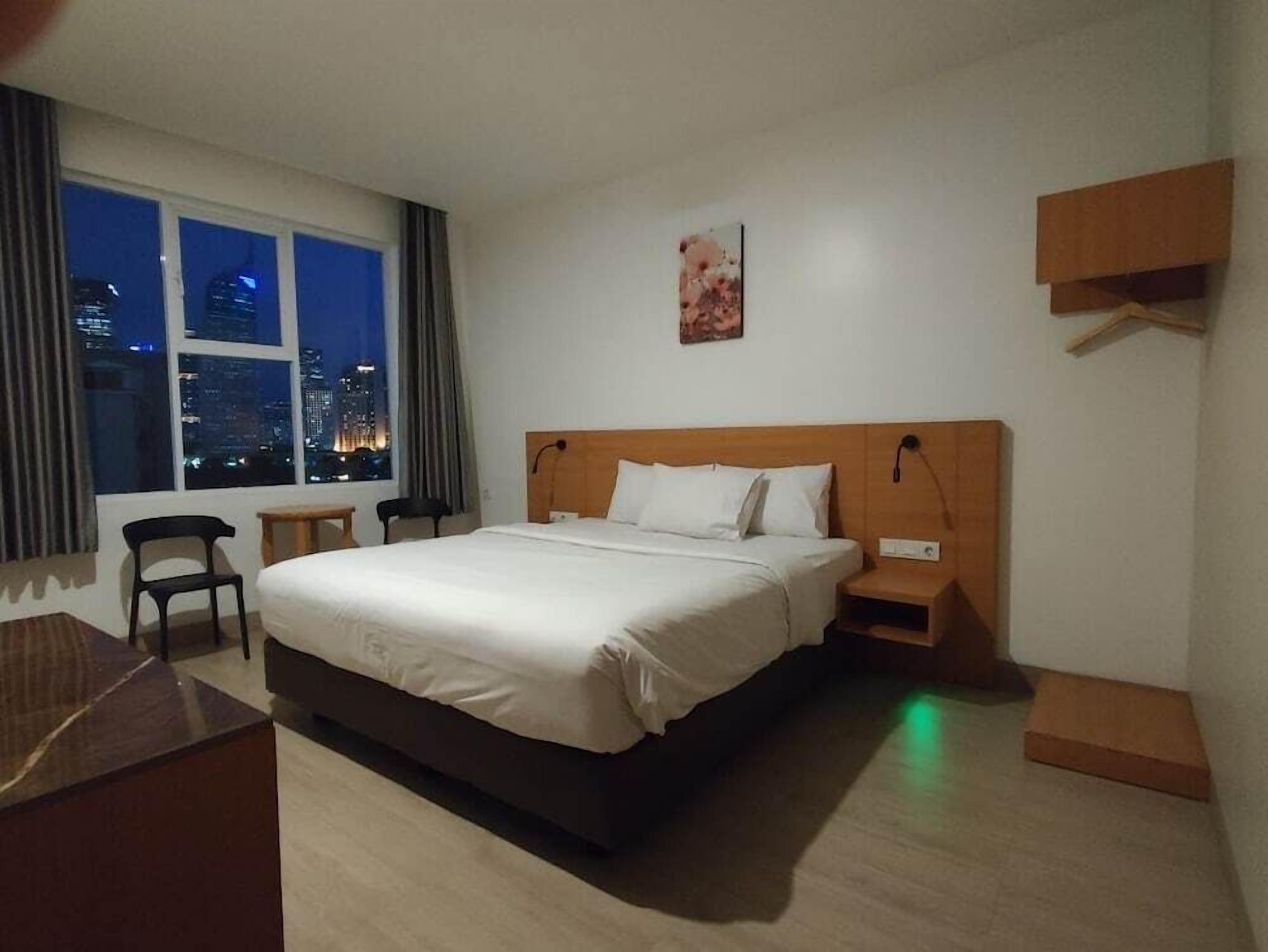 Une chambre d’hôtel avec un grand lit, un bureau, une chaise, une table de chevet et une vue sur le paysage urbain la nuit.