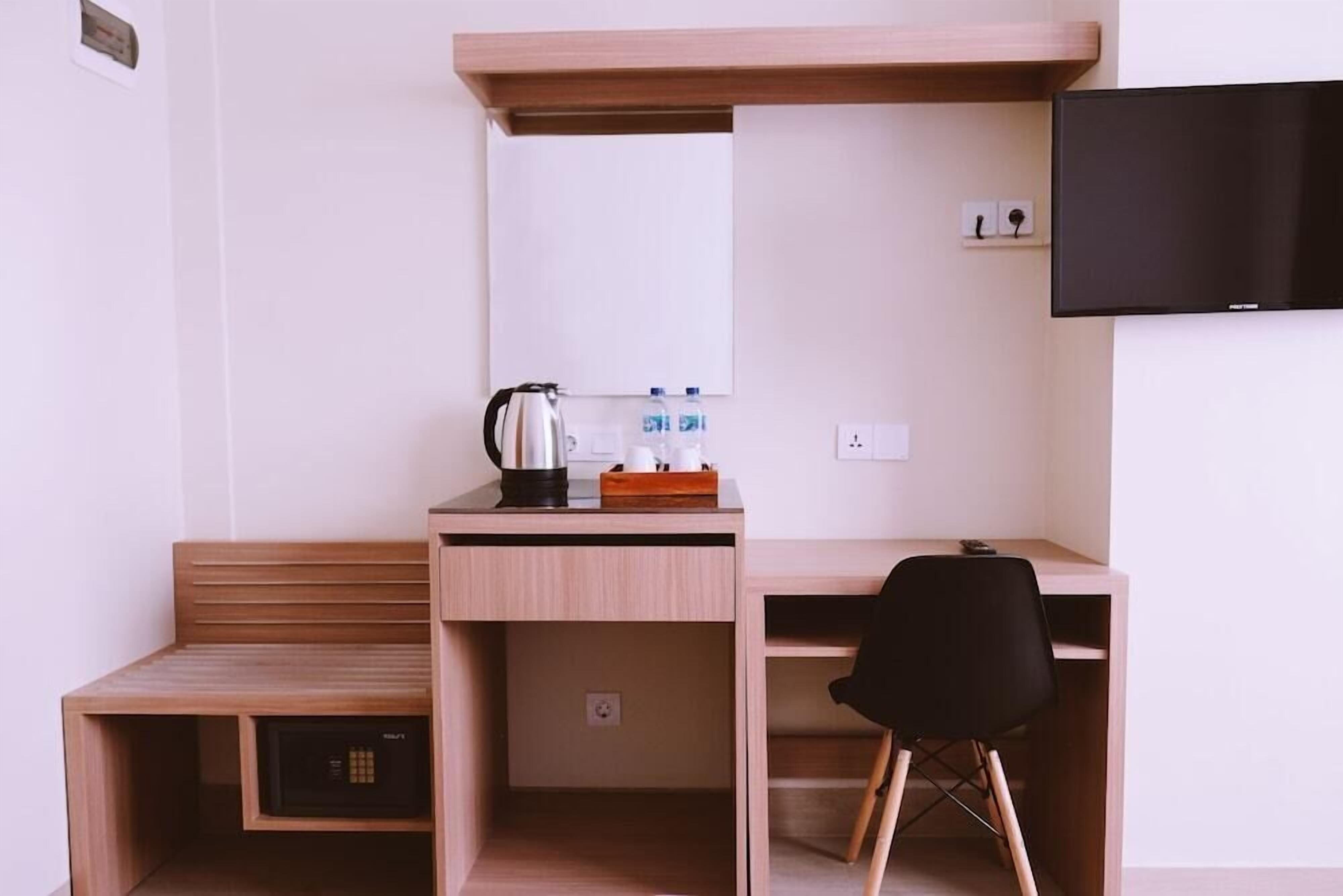 A compact, modern room with a wooden desk, a black chair, a small heater, a kettle, and a flat-screen TV mounted on the wall.