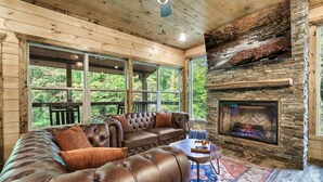A cozy living room with a leather sectional sofa, a stone fireplace, and large windows offering a view of the outdoors. - Best Family Cabin - Indoor Pool, Cinema, Arcade, Putt Putt, Spa, Fire Pit, Creek (Sevierville)
