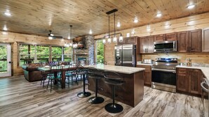 A modern kitchen with wooden cabinets, a central island with bar stools, a stone fireplace, and a large window overlooking greenery. - Best Family Cabin - Indoor Pool, Cinema, Arcade, Putt Putt, Spa, Fire Pit, Creek (Sevierville)
