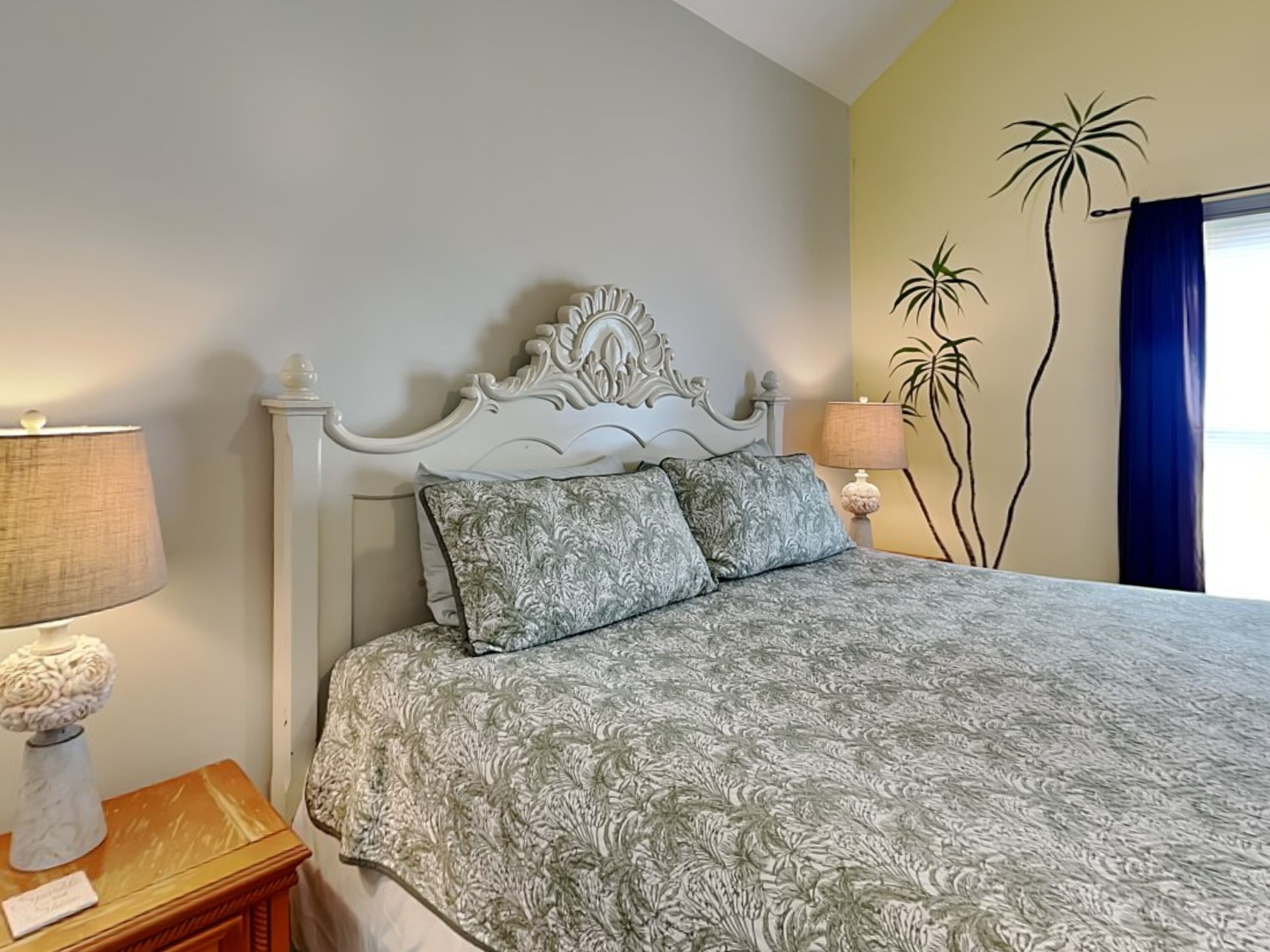 A neatly made bed with a patterned bedspread, a white ornate headboard, and a nightstand with a lamp and a decorative vase.