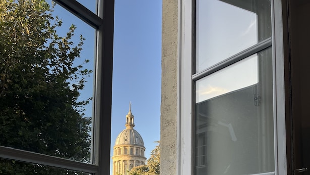 Une fenêtre donnant sur un grand bâtiment à coupole, avec un rebord de fenêtre blanc et une rambarde métallique à l’extérieur.